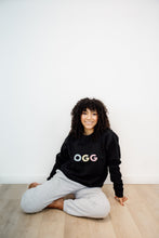 Load image into Gallery viewer, woman wearing black crewneck with the OGG airport code written on the front in colourful graphics
